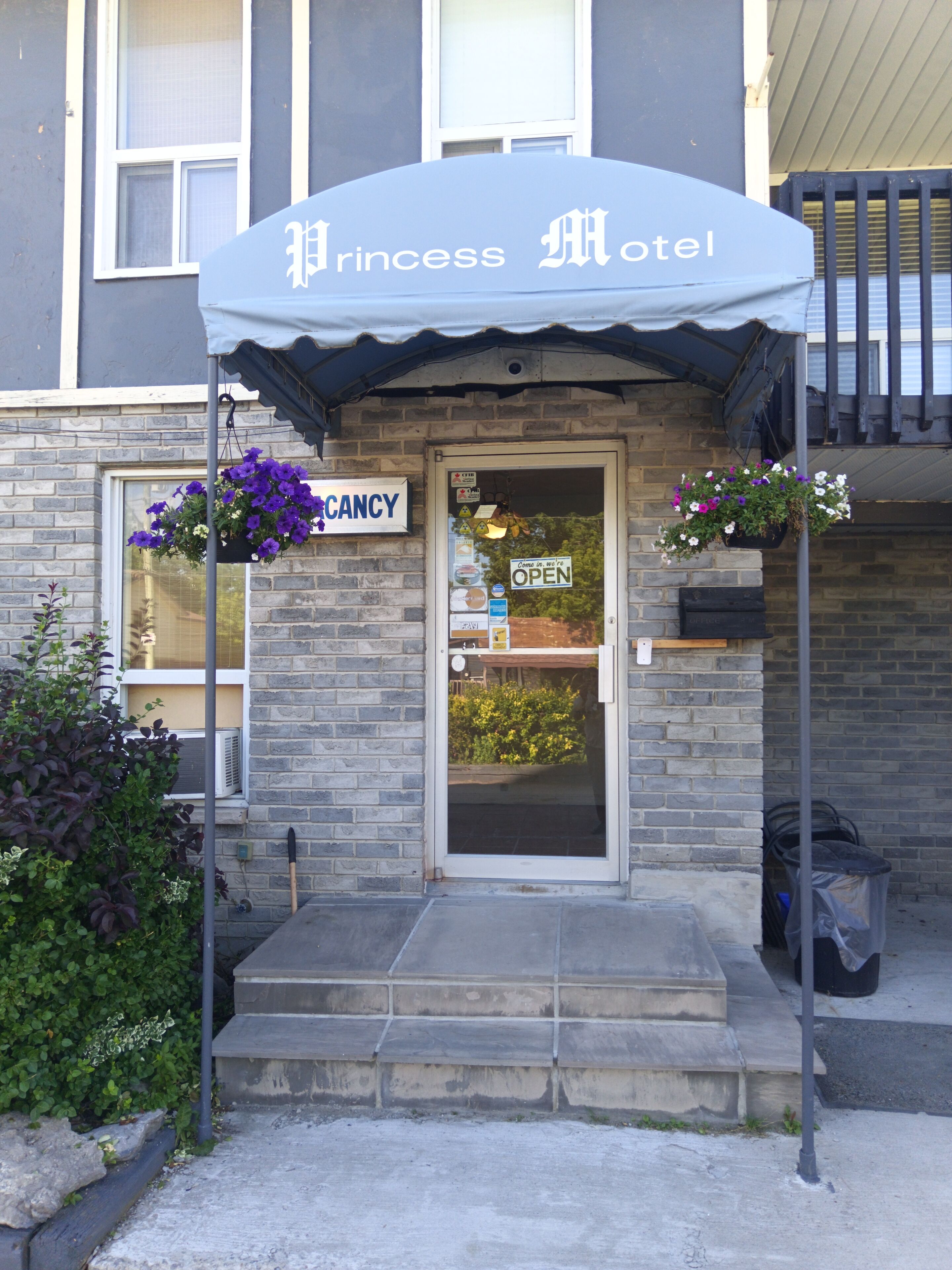 Entrada de un hotel con un cartel que dice "Princess Hotel" y un letrero "CANCY". Hay cestos de flores colgando en la pared y un bote de basura afuera.