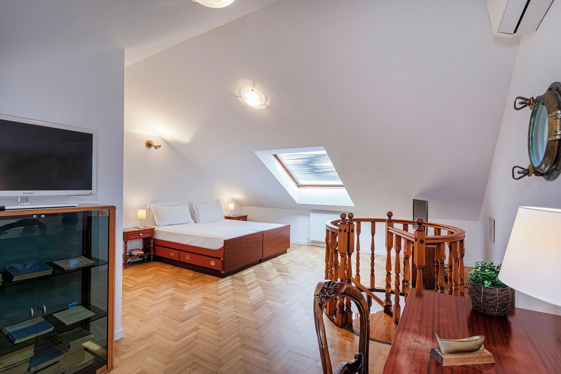 A bedroom with a bed, bedside tables, a TV, a skylight, a wooden chair, and a bookshelf.