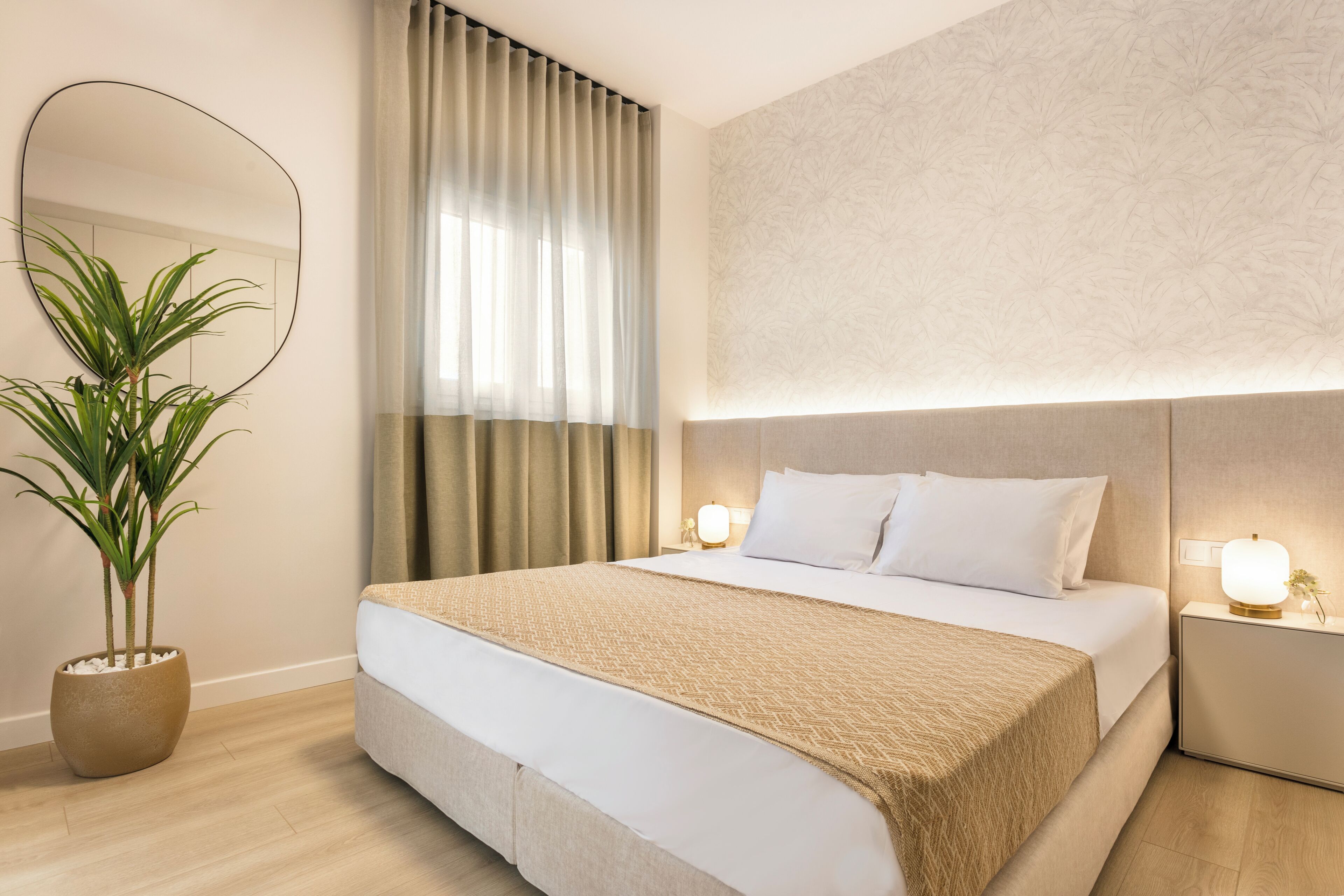 A modern hotel room with a large bed, bedside lamps, a potted plant, and a large mirror.