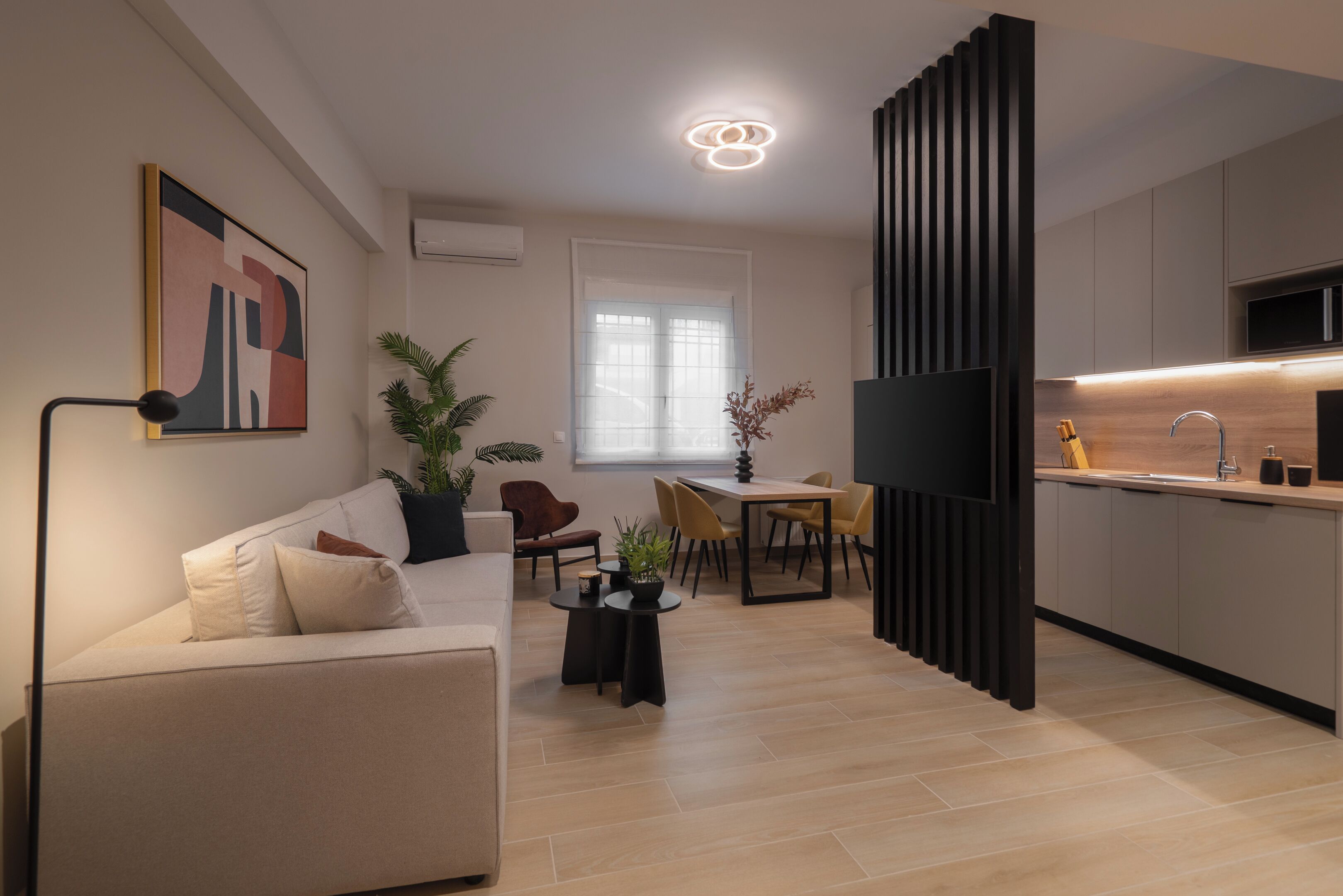 A modern living room with a sofa, a dining table, and chairs. There is a TV mounted on the wall and a kitchen area with a sink and cabinets.
