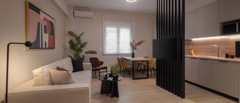 A modern living room with a sofa, a dining table, and chairs. There is a TV mounted on the wall and a kitchen area with a sink and cabinets.