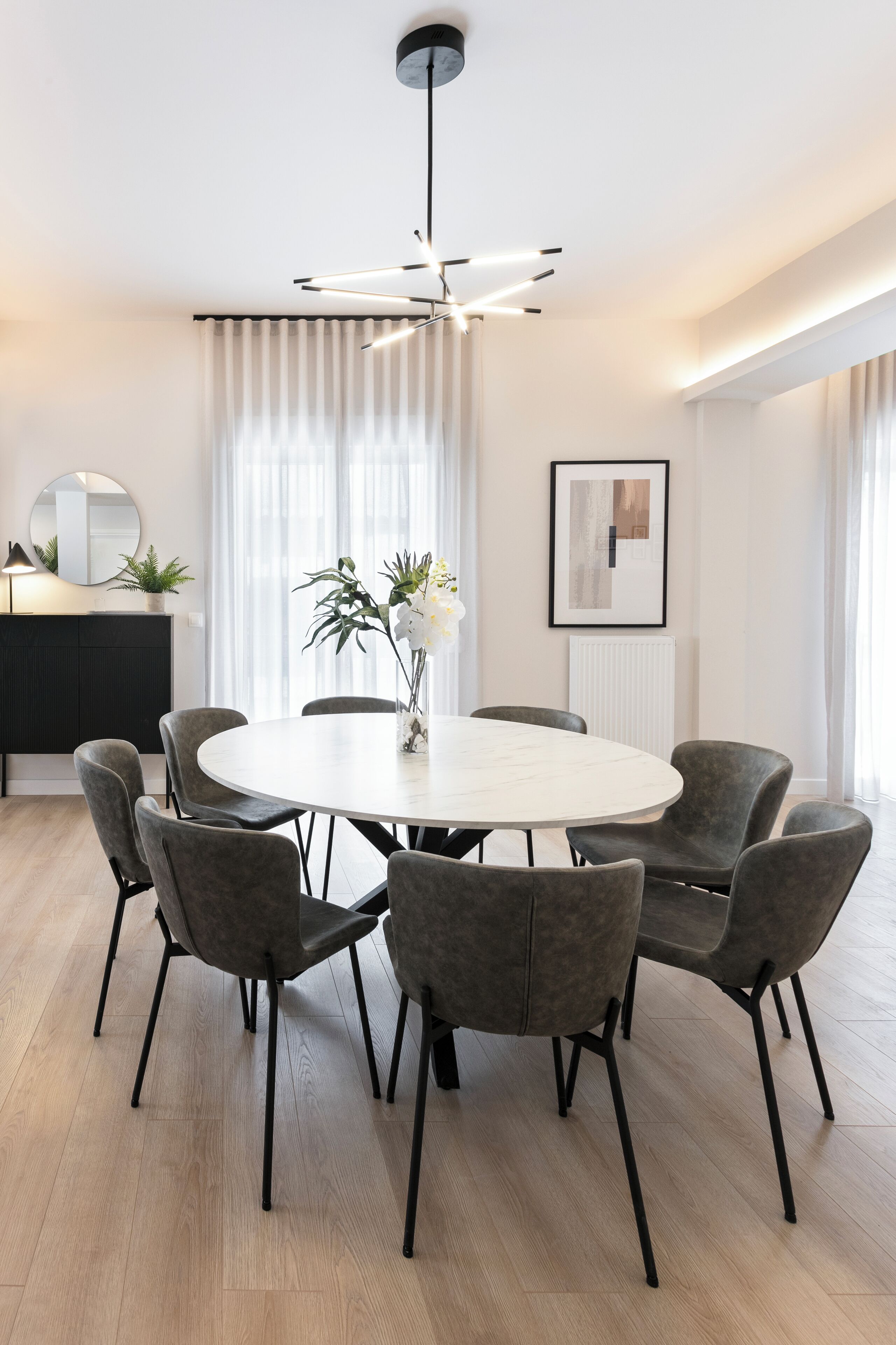 A modern dining area with a round table, six grey chairs, a pendant light, a potted plant, and a framed artwork on the wall.