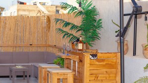 A wooden outdoor seating area with a table and chairs, a bar counter with a bottle and glasses, and a large umbrella providing shade. - Le Rooftop Céleste Apparte (Nabeul‎)