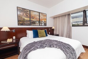 A hotel room with a bed, a nightstand with a phone, a window with curtains, and a wall with three paintings.