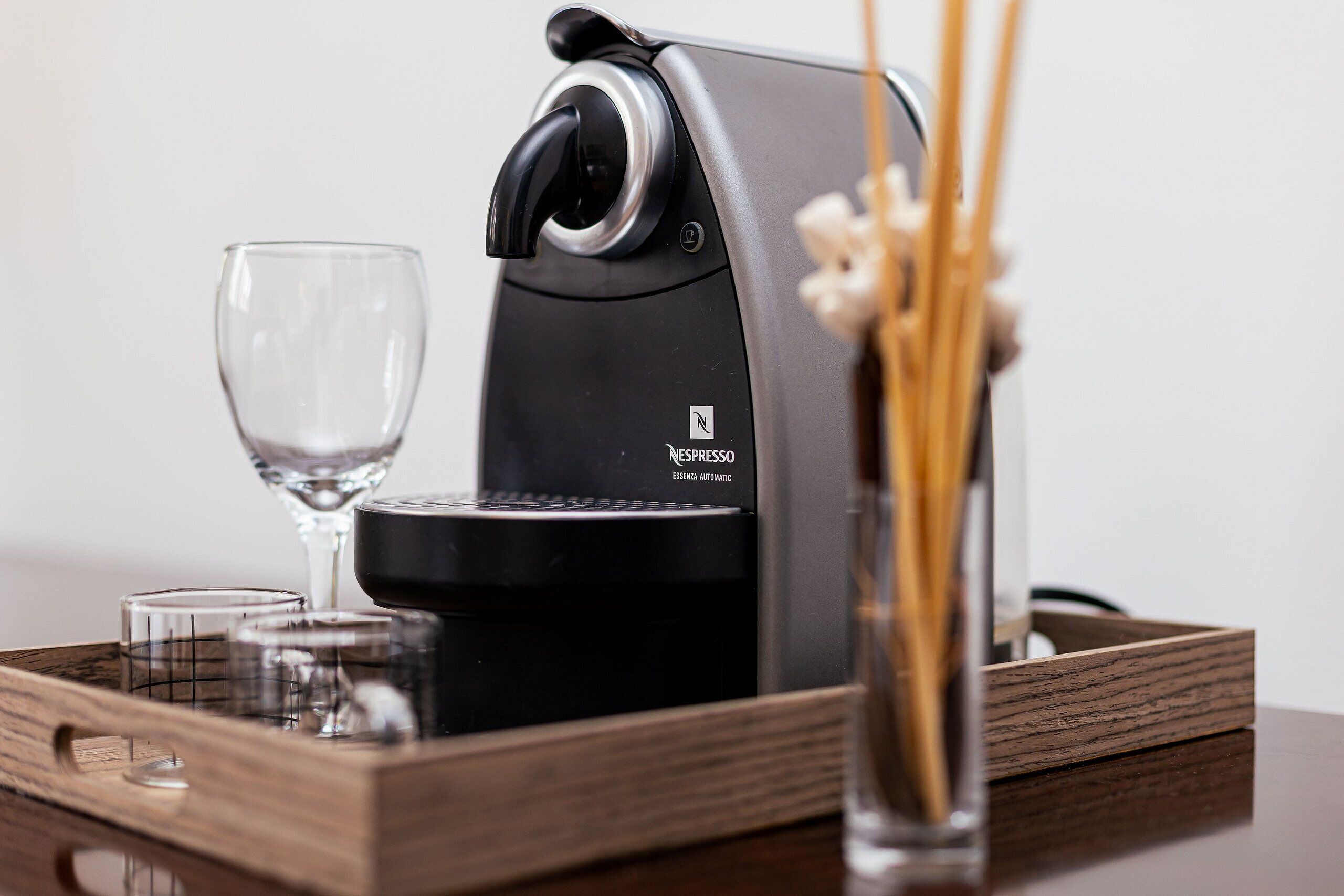 A Nespresso espresso machine on a wooden tray with two glasses and a vase with reeds.