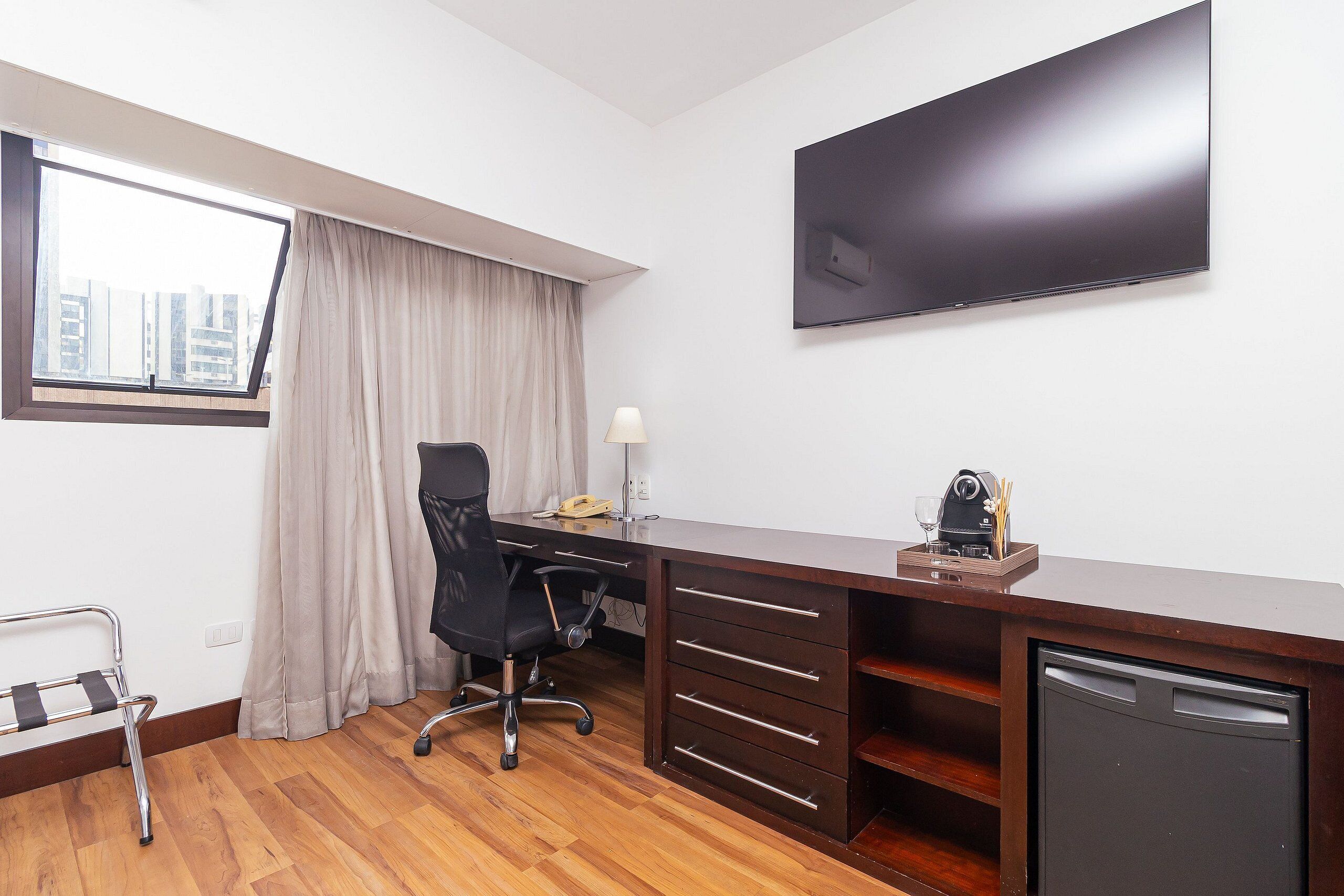 Hotelkamer met een houten bureau, een stoel, een aan de muur bevestigde televisie, een raam met gordijnen en een kleine koelkast.