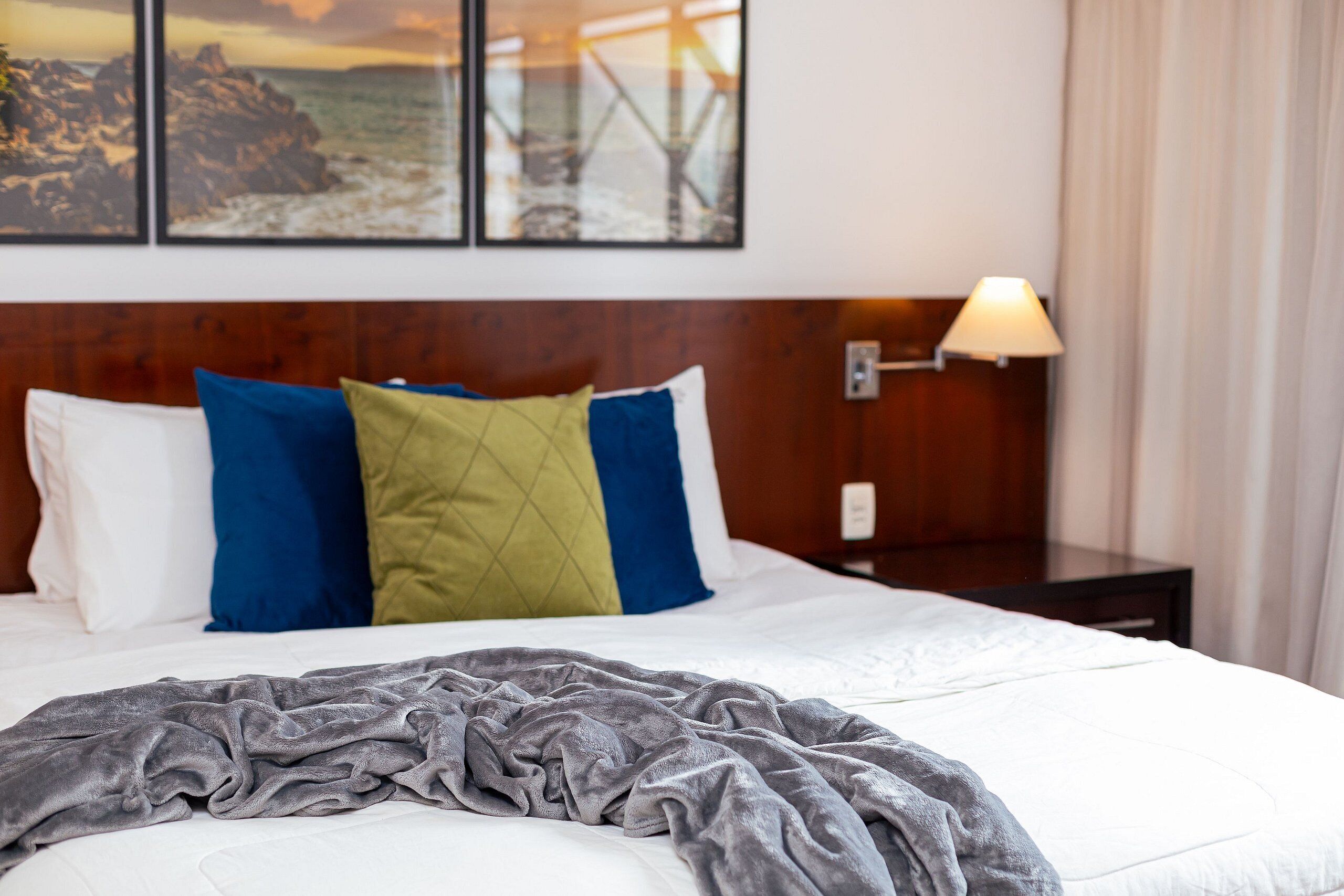 Cama bem arrumada com manta cinzenta, travesseiros brancos e um travesseiro azul e verde. Uma luminária está sobre a mesa de cabeceira, e uma moldura com uma imagem de paisagem costeira está na parede.