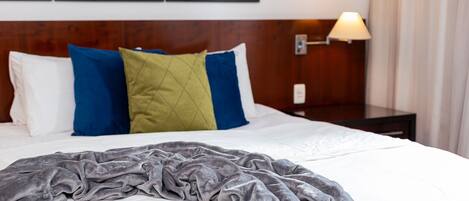 Cama bem arrumada com manta cinzenta, travesseiros brancos e um travesseiro azul e verde. Uma luminária está sobre a mesa de cabeceira, e uma moldura com uma imagem de paisagem costeira está na parede.