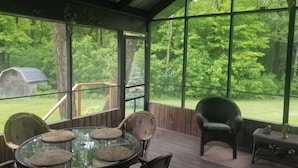A covered outdoor area with a glass table and chairs, surrounded by large windows offering a view of a wooded area. - Wagonwheel Ranch (Viroqua)