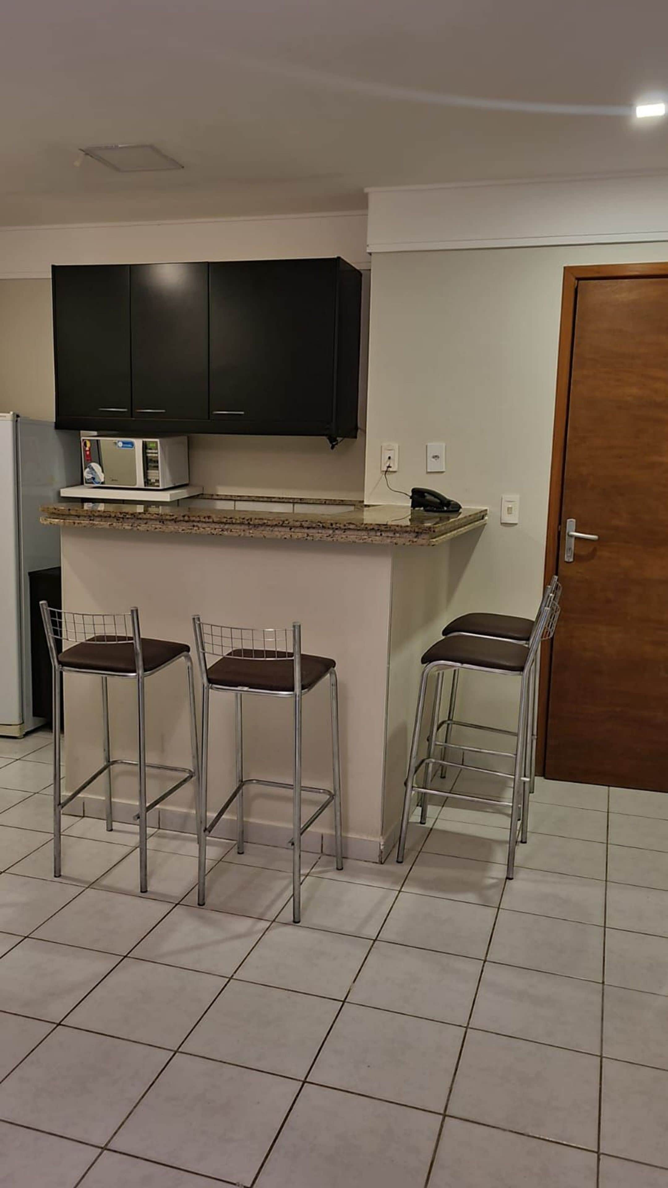 Una cocina con armarios oscuros, un microondas y un refrigerador pequeño. Hay tres sillas metálicas con cojines. Se ve una puerta de madera al fondo.