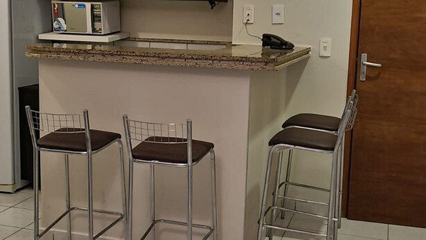 Eine Küche mit dunklen Schränken, einer Mikrowelle und einem kleinen Kühlschrank. Drei Metallstühle mit Sitzkissen sind vorhanden. Im Hintergrund ist eine Holztür zu sehen.