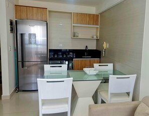 A compact kitchen with a refrigerator, stove, and microwave. There is a glass table with chairs and a small cabinet above. - CJC Short Stay Natal (Natal)
