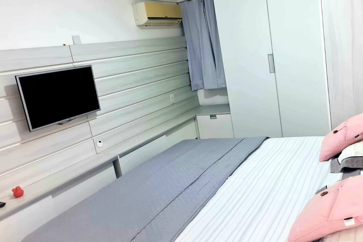 Et hotelværelse med en seng, et fjernsyn ophængt på væggen, et klædeskab og et lille bord med en fjernbetjening.