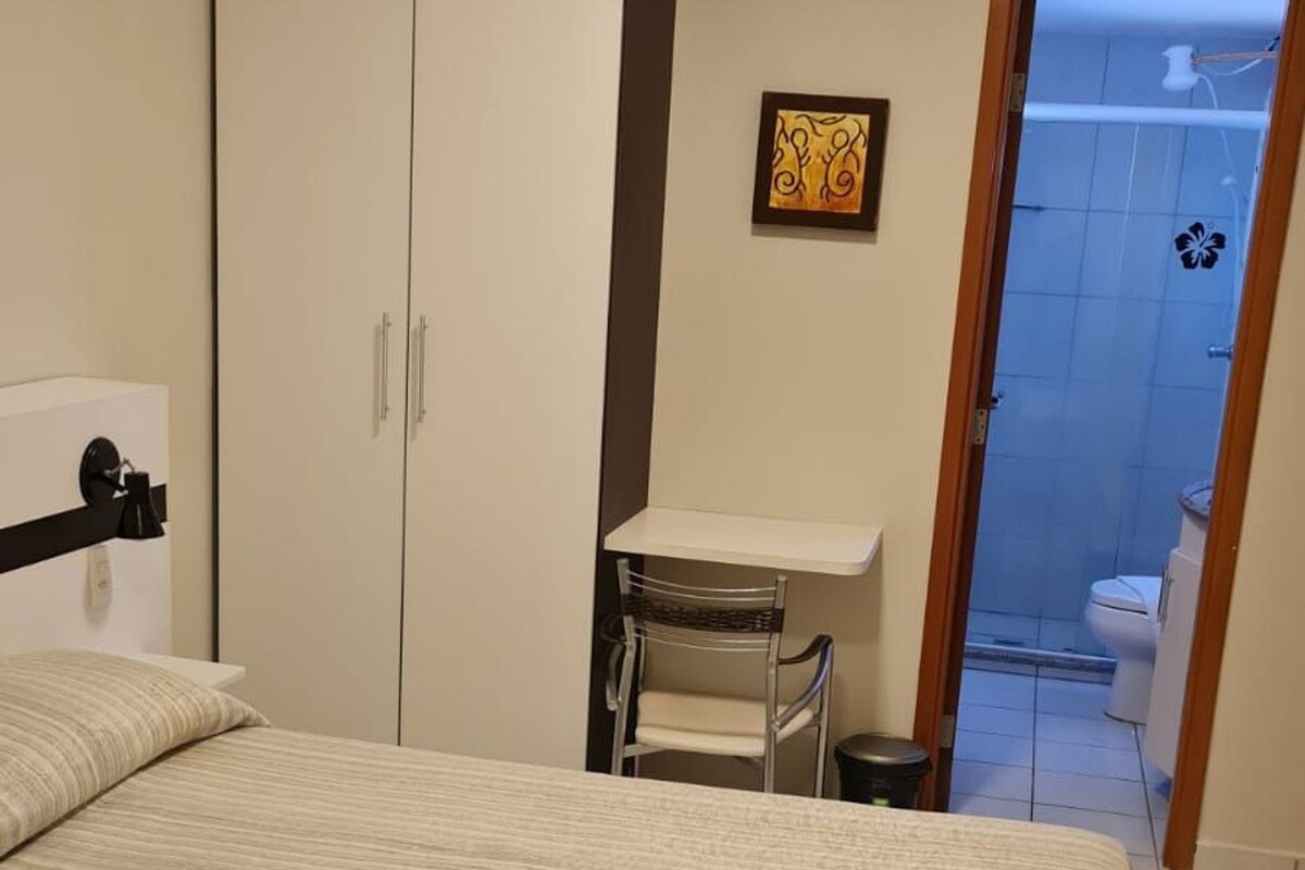 A bedroom with a bed, a desk, and a chair. There is a bathroom visible through an open door.