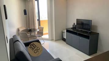 Uma sala de estar moderna com um sofá, uma mesa de jantar com cadeiras, uma televisão sobre um móvel e uma janela grande com cortinas.