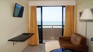 A hotel room with a sofa, a small table, a TV mounted on the wall, and a view of the sea through a sliding door. - CJC Short Stay Natal (Natal)