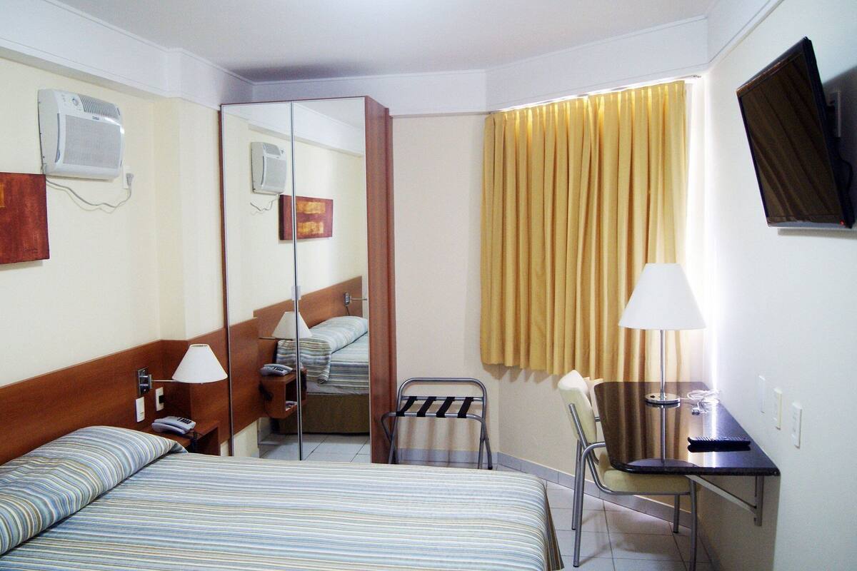 Et hotelværelse med en seng, sengeborde med lamper, et skrivebord med stol, et fjernsyn og et spejl.