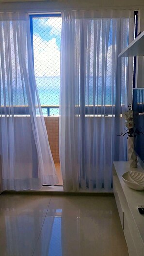 A room with a view through sheer curtains to a balcony, a white vase, and a bowl on a white console table. - CJC Short Stay Natal (Natal)