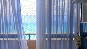 A room with a view through sheer curtains to a balcony, a white vase, and a bowl on a white console table. - CJC Short Stay Natal (Natal)