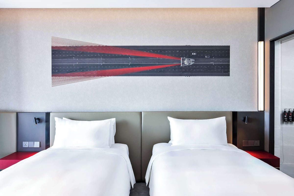 Ein Hotelzimmer mit zwei Betten, einem modernen Kopfteil, einem an der Wand befestigten Kunstwerk und einem Nachttisch mit roter Oberfläche.