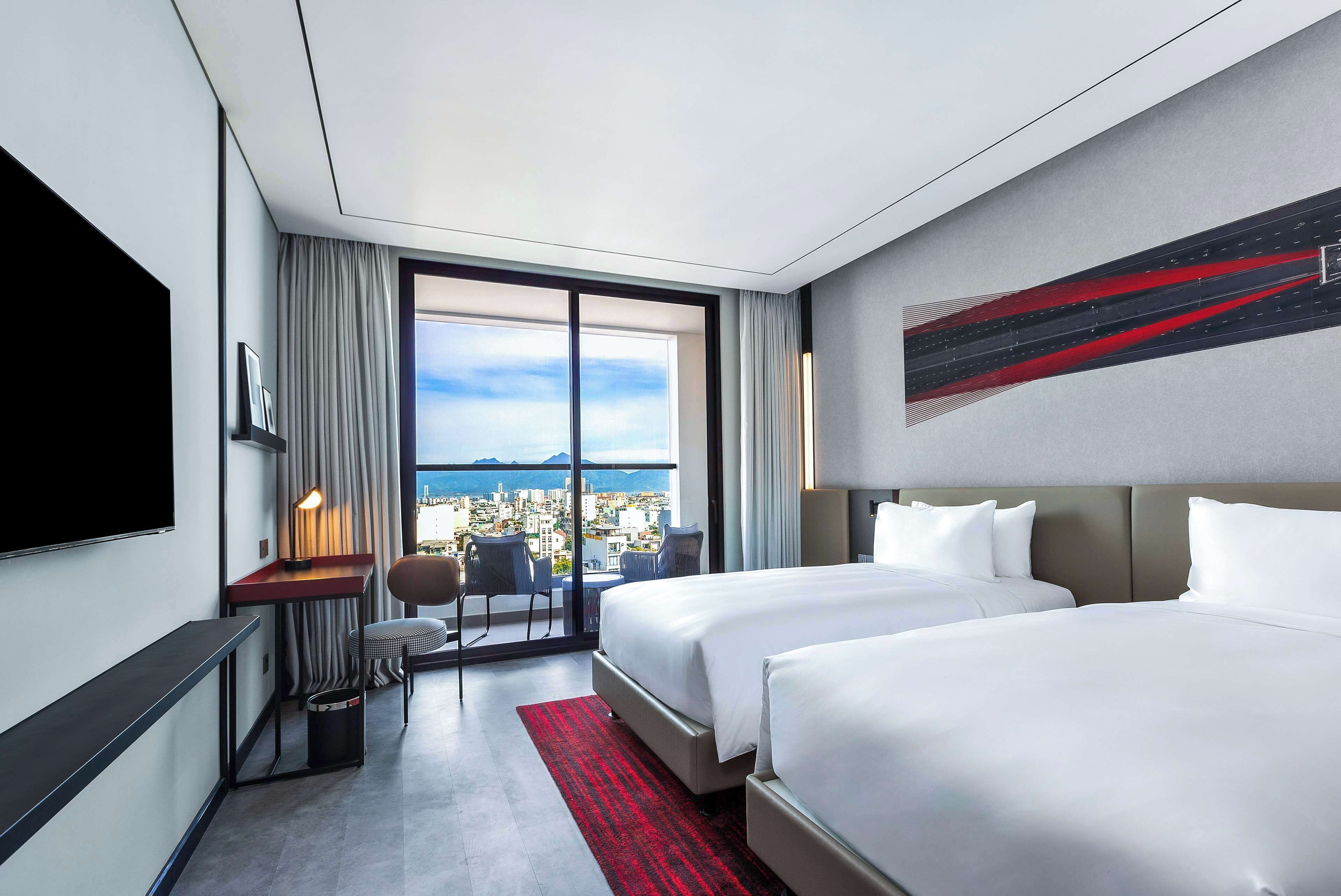 A hotel room with two beds, a desk, a chair, a TV, and a large window with a city view.