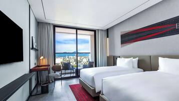 A hotel room with two beds, a desk, a chair, a TV, and a large window with a city view.