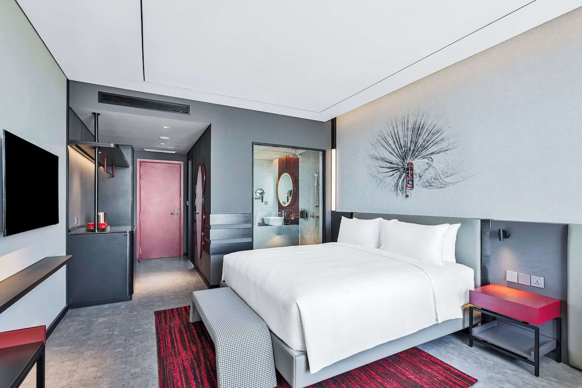 Ein modernes Hotelzimmer mit einem großen Bett, einem Fernseher, einem roten Nachttisch und einem Badezimmer mit Spiegel und Waschbecken.