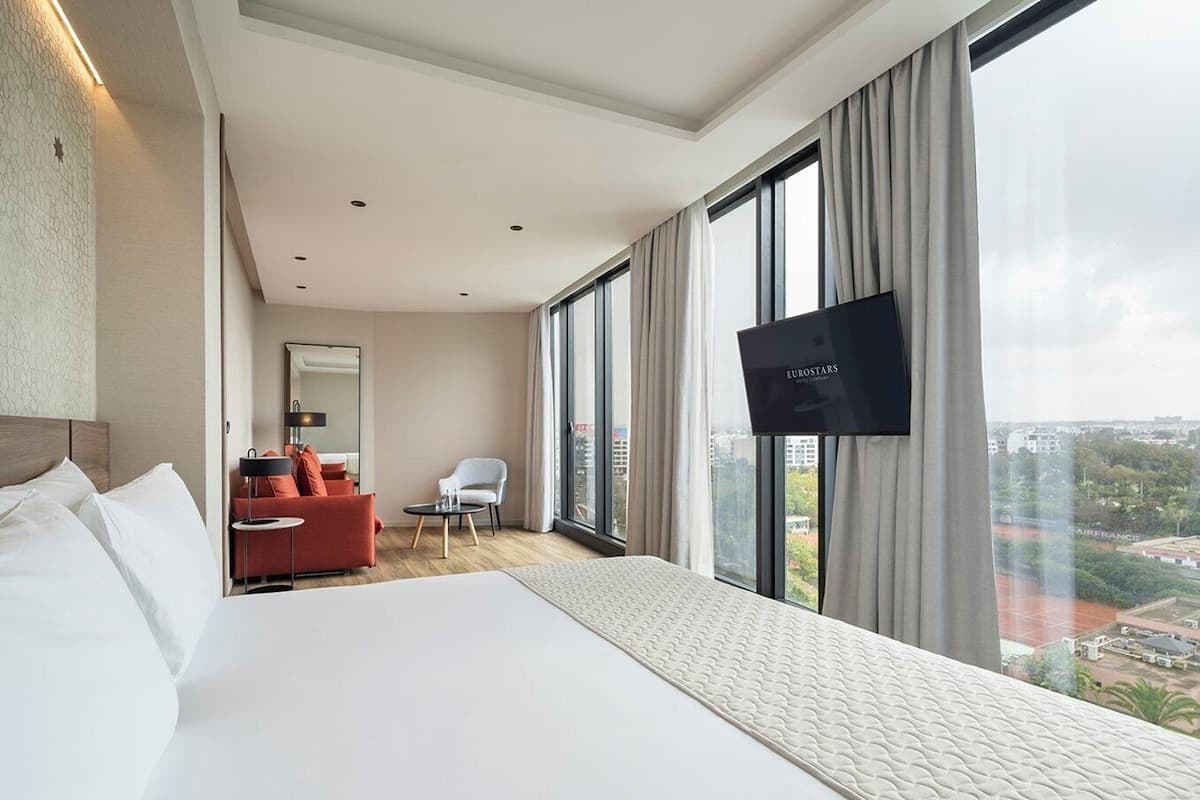 Une chambre d’hôtel moderne dotée d’un grand lit, d’un téléviseur fixé au mur, d’un coin salon avec un canapé rouge et un fauteuil, et d’une grande fenêtre donnant sur la ville.