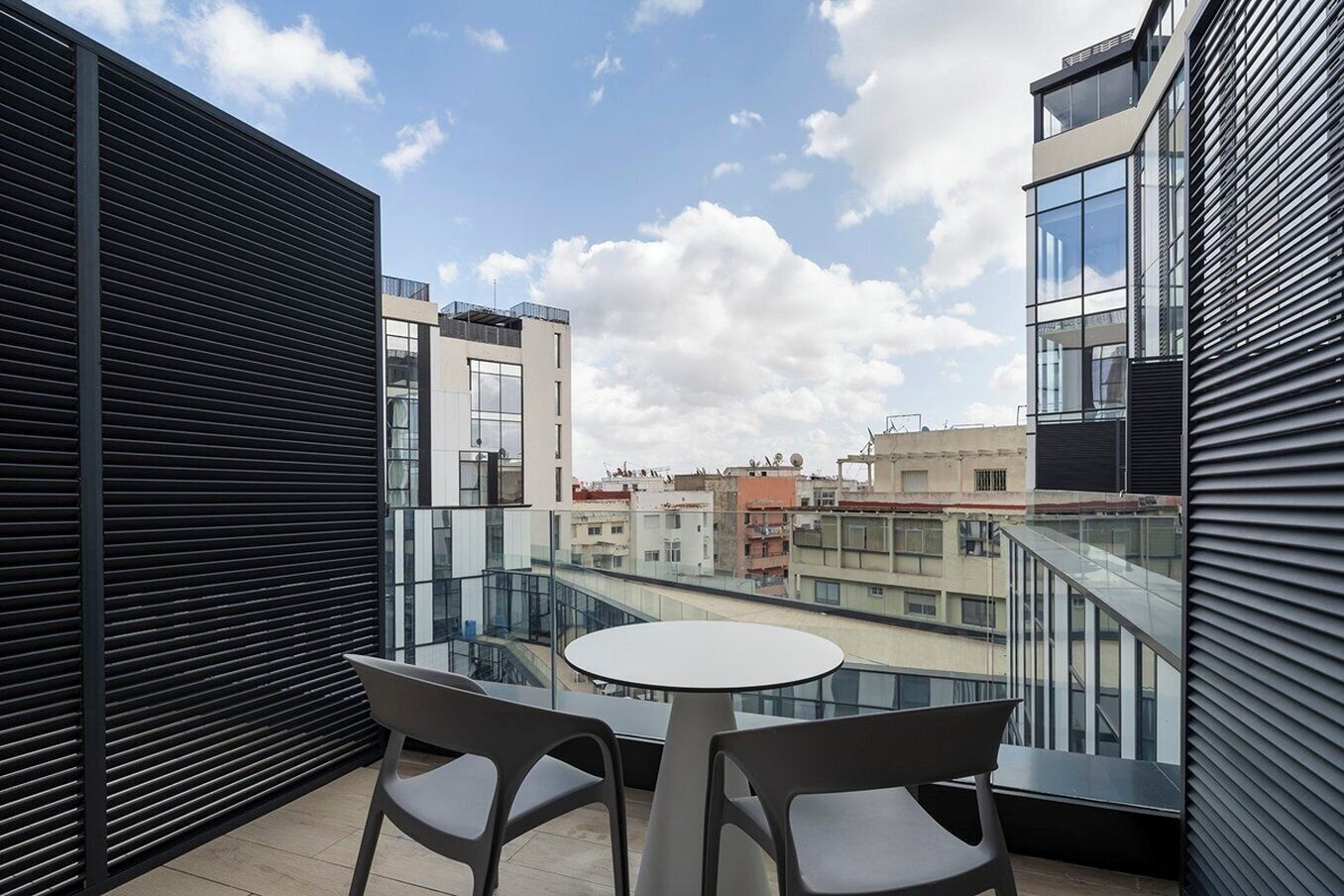A balcony with two chairs and a table, overlooking a cityscape with various buildings.