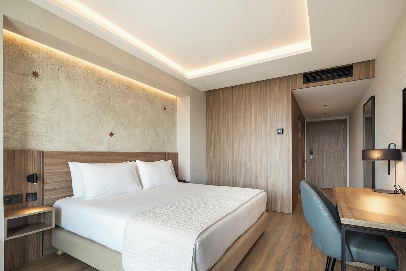 A modern hotel room with a large bed, a desk, and a built-in wardrobe.
