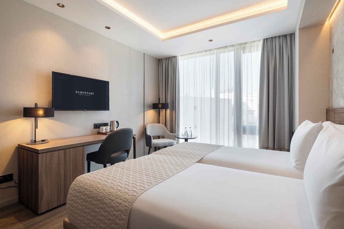 A hotel room with a bed, a desk with a chair, a TV, and a window with curtains.