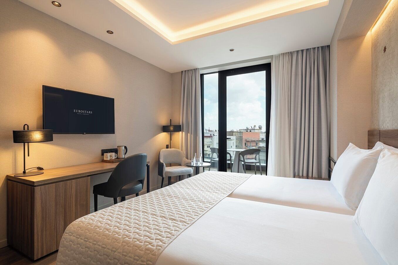 Habitación de hotel con cama, escritorio, sillas, televisor y vista a la ciudad.