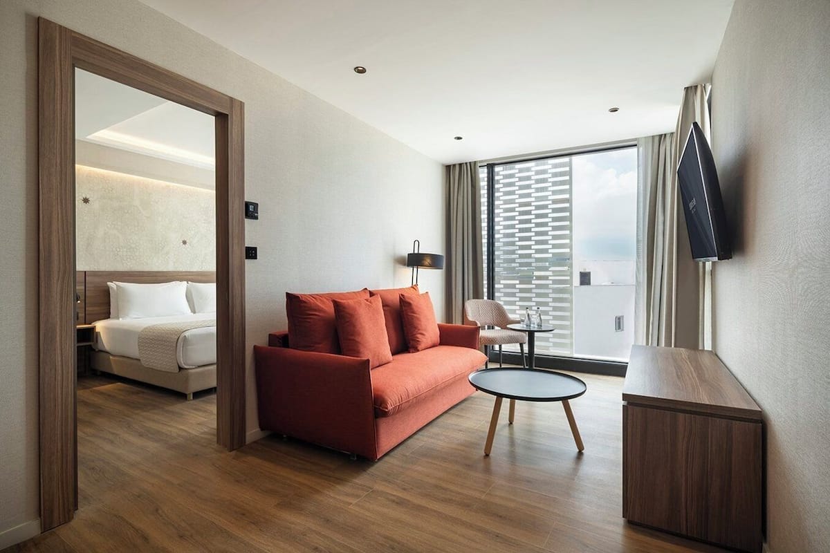 Une chambre d’hôtel moderne avec un plancher en bois, un canapé rouge, une petite table ronde, un téléviseur fixé au mur et un balcon avec vue.