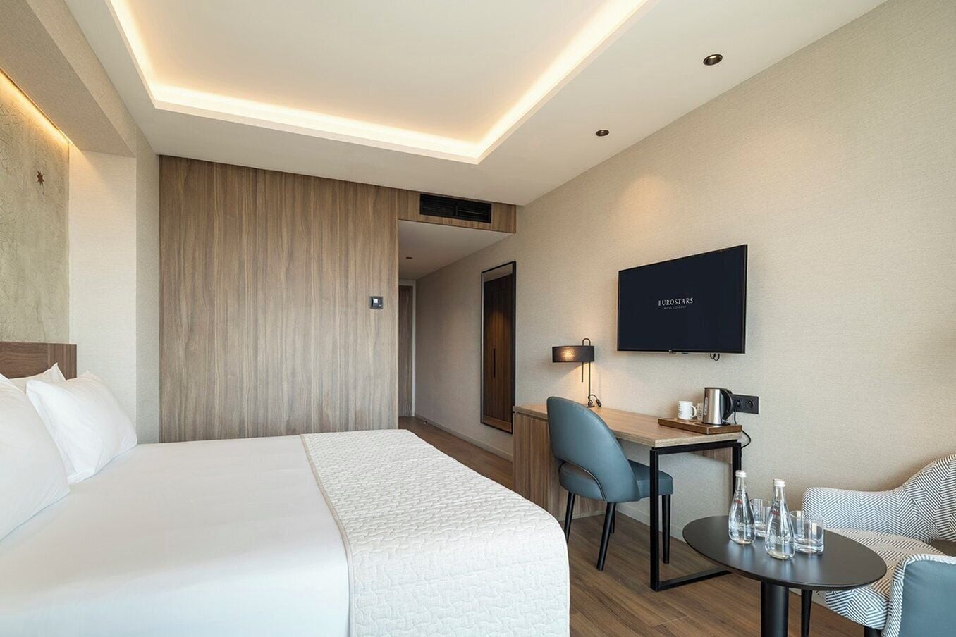 A hotel room with a bed, a desk with a chair, a TV, and a small table with glasses and a bottle.