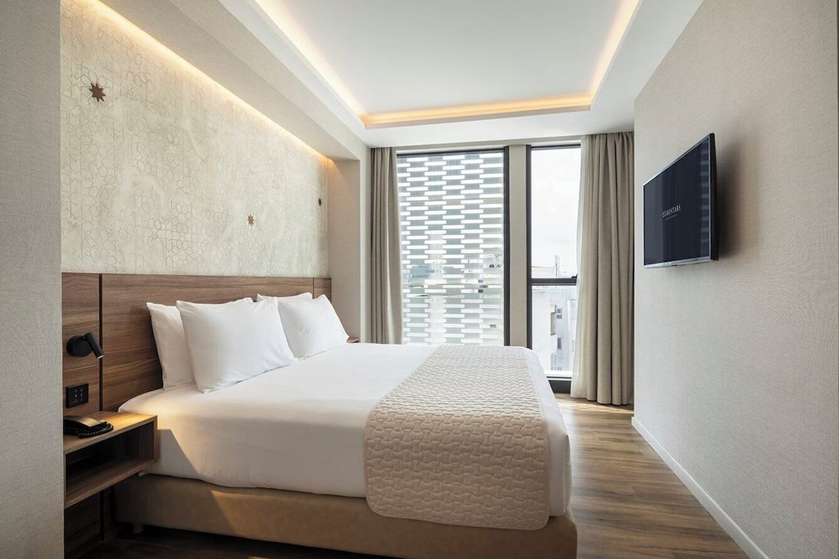 Une chambre d’hôtel moderne dotée d’un grand lit, d’une table de chevet avec une lampe, d’un téléviseur à écran plat fixé au mur et offrant une vue sur la ville grâce à de grandes fenêtres.