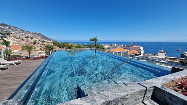 Ein rechteckiger Infinity-Pool mit klarem, blauem Wasser, umgeben von einer Holzterrasse und einer Steinbordsteinkante. Im Hintergrund ist ein Blick auf eine Küstenstadt mit Gebäuden und einem strahlend blauen Himmel zu sehen.
