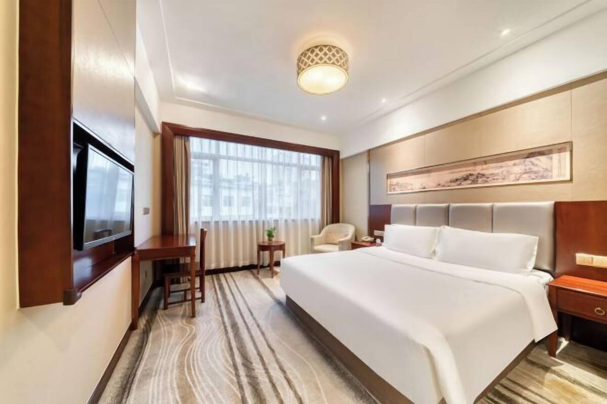 Une chambre d’hôtel avec un grand lit, un bureau, une télévision, une fenêtre avec des rideaux et un tableau au mur.