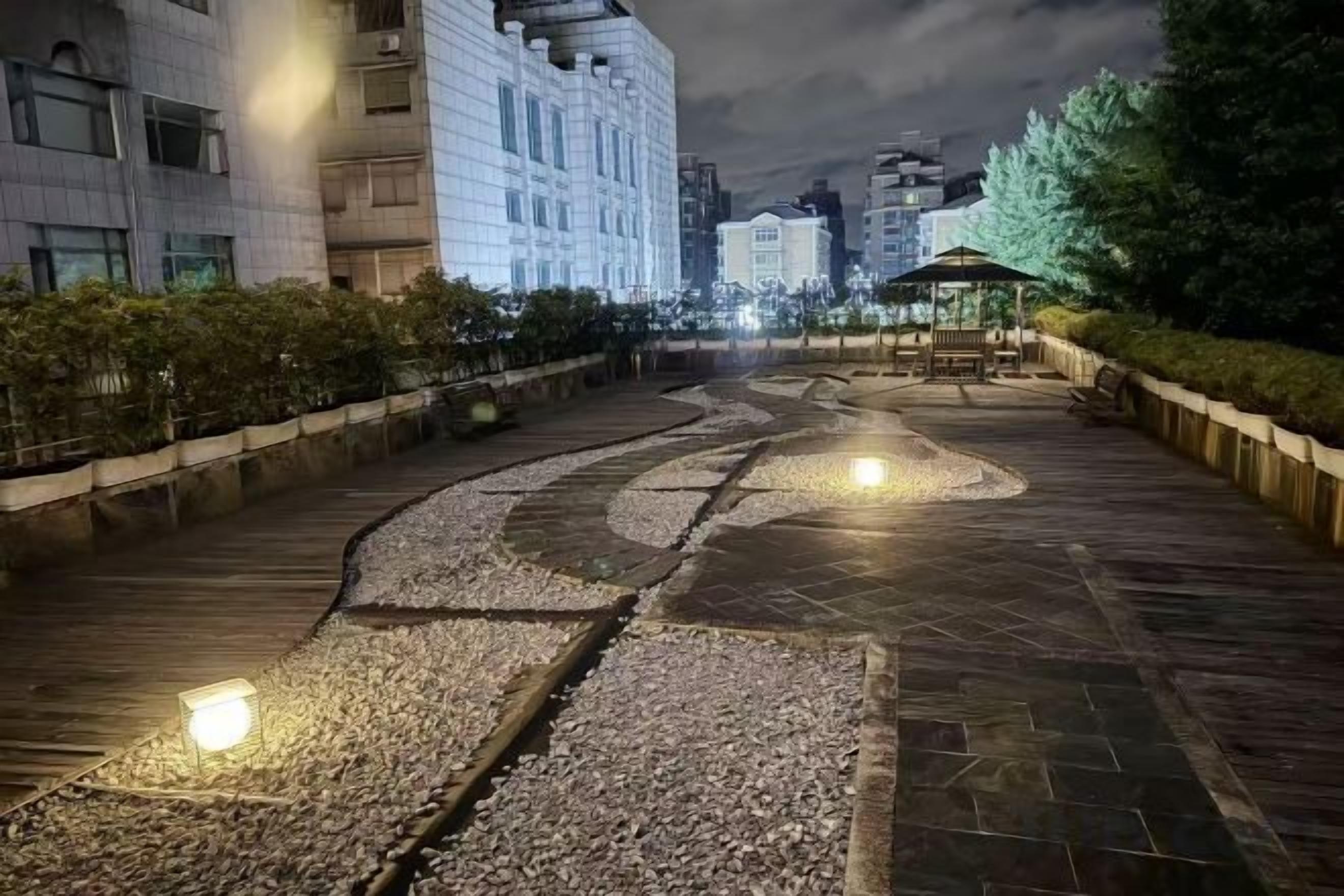 A paved pathway with a gentle curve, flanked by buildings and greenery, leading to a gazebo.