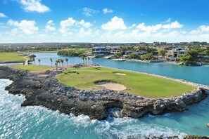 A coastal golf course with a water body, rocky shoreline, and buildings in the background.