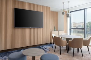 A modern conference room with a large flat-screen TV, a round table with chairs, and a large window offering a view of the outside. - Courtyard Milano Linate (Milan)