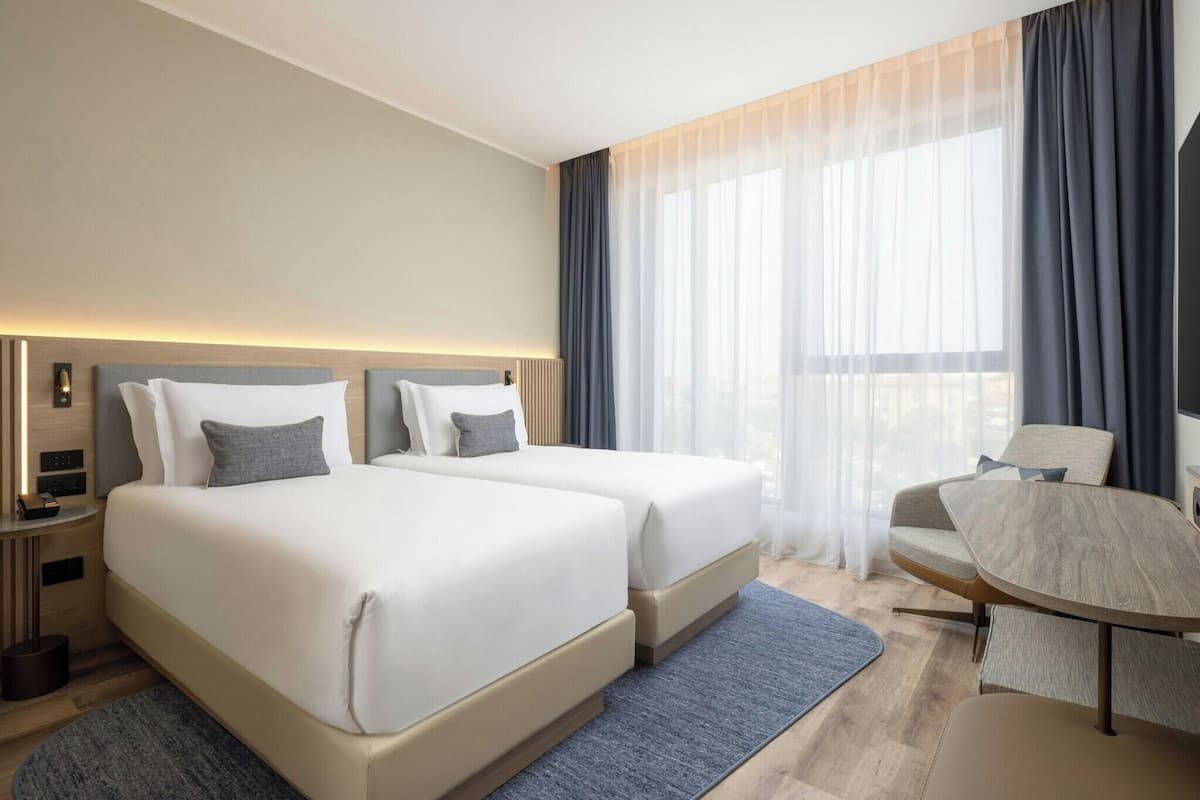 A modern hotel room with two beds, a desk, and a chair.