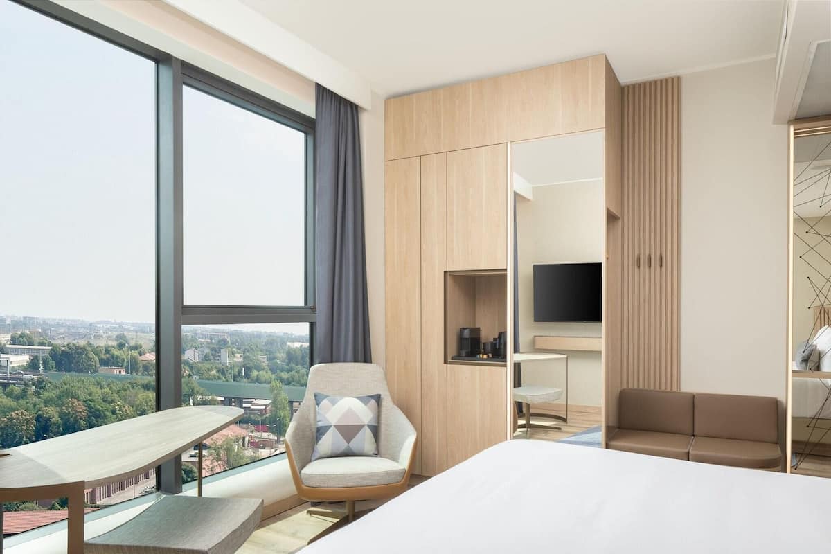 A modern hotel room with a large window, a bed, a desk, a chair, a sofa, and a TV.