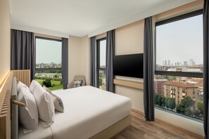 Ein modernes Hotelzimmer mit einem großen Bett, einem Fernseher und Blick auf die Stadt durch große Fenster.