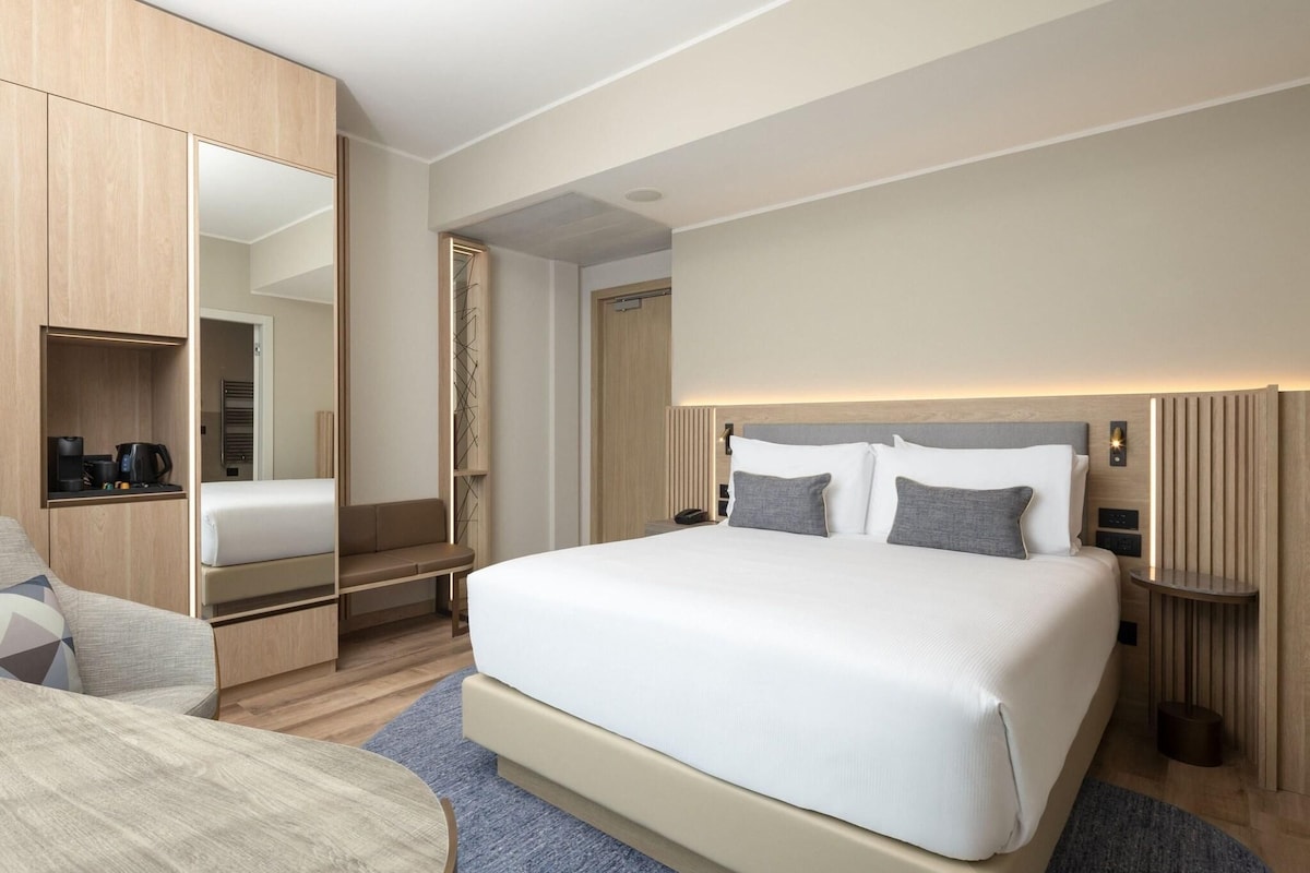 A modern hotel room with a large bed, a built-in wardrobe, a seating area, and a small table.