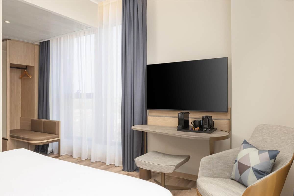 A hotel room with a bed, a seating area with a chair and a small table, a TV mounted on the wall, and a wardrobe.