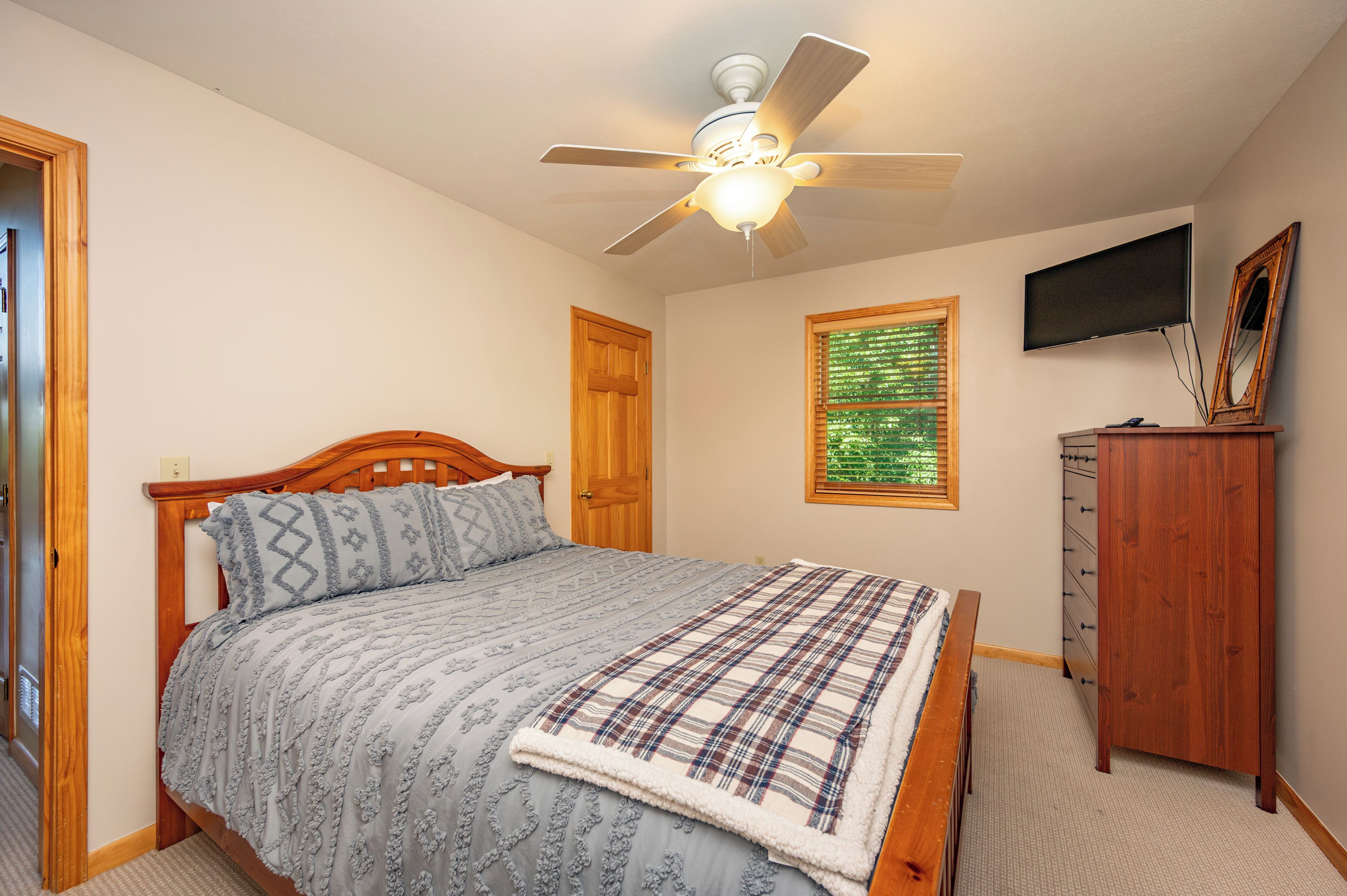 A bedroom with a bed, a ceiling fan, a television, a mirror, and a window with blinds.