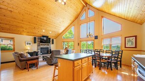A spacious living area with a wooden ceiling, a fireplace, a dining table with chairs, and large windows offering a view of the outdoors. - Scenic Retreat w/ Pool Table & Lake Views (Swanton)