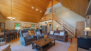 A spacious living area with a wooden ceiling, a dining area with chairs and a table, a kitchen with cabinets and appliances, and a staircase leading to an upper level. - Scenic Retreat w/ Pool Table & Lake Views (Swanton)