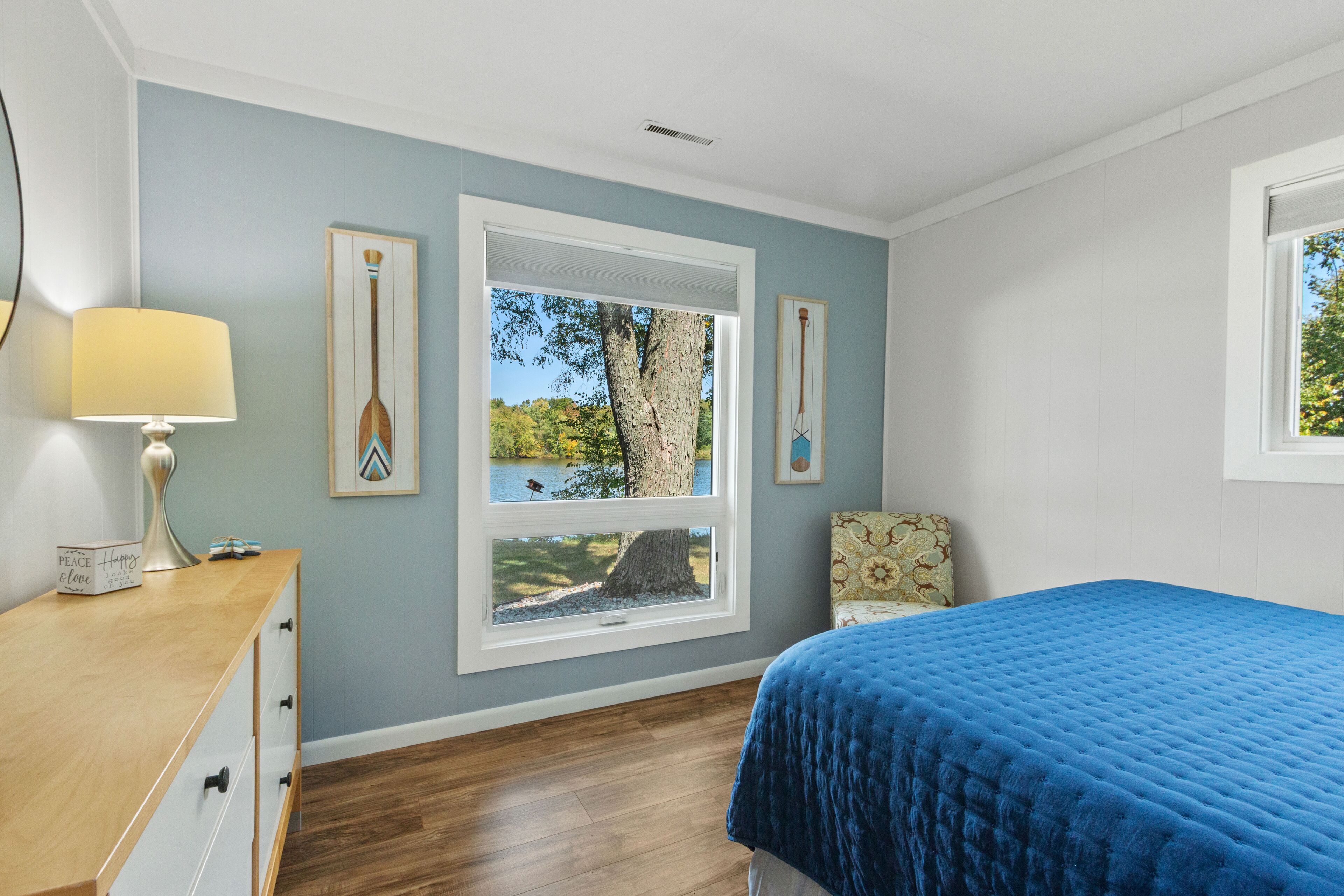 A bedroom with a bed, a desk, a chair, and a window with a view of trees and a lake.