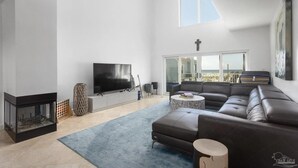 A modern living room with a fireplace, a large flat-screen TV, a sectional sofa, and a view of the outdoors through a large window. - Portside Villas Unit 6 (Pensacola Beach)